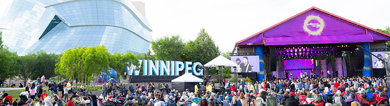 a crowd of people seated in lawn chairs watching a performance on a stage. A large Winnipeg sign is in the background in front of the Human Rights Museum.